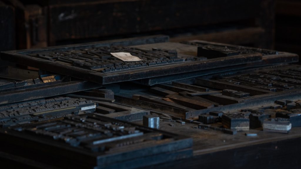 Stacked letterpress formes of metal type on a workbench in a dim print workshop.