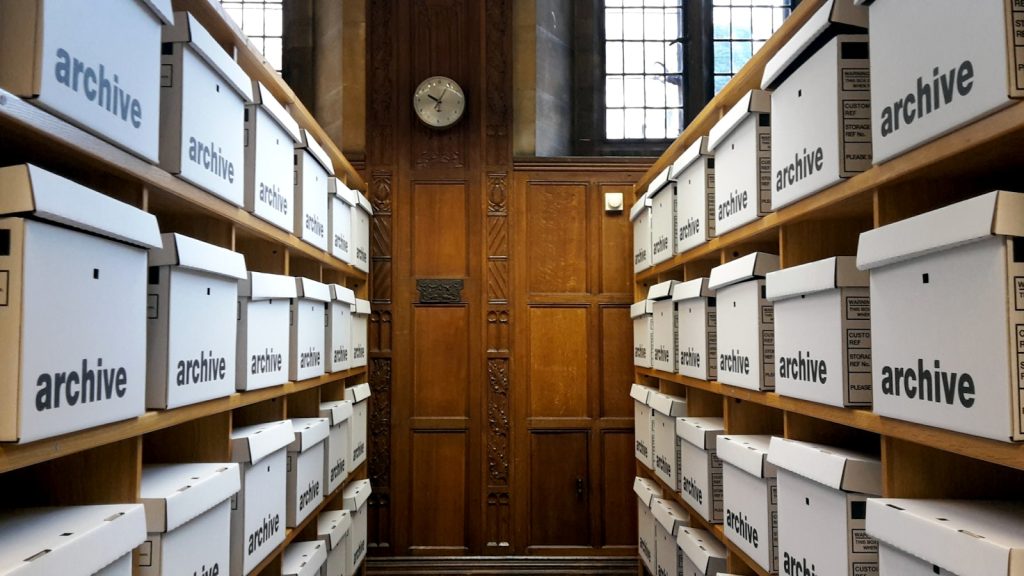 Rows of white archive boxes stacked on wooden shelving in a wood-panelled room.