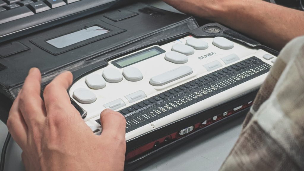 A person's hands reading a refreshable braille display connected to a keyboard.